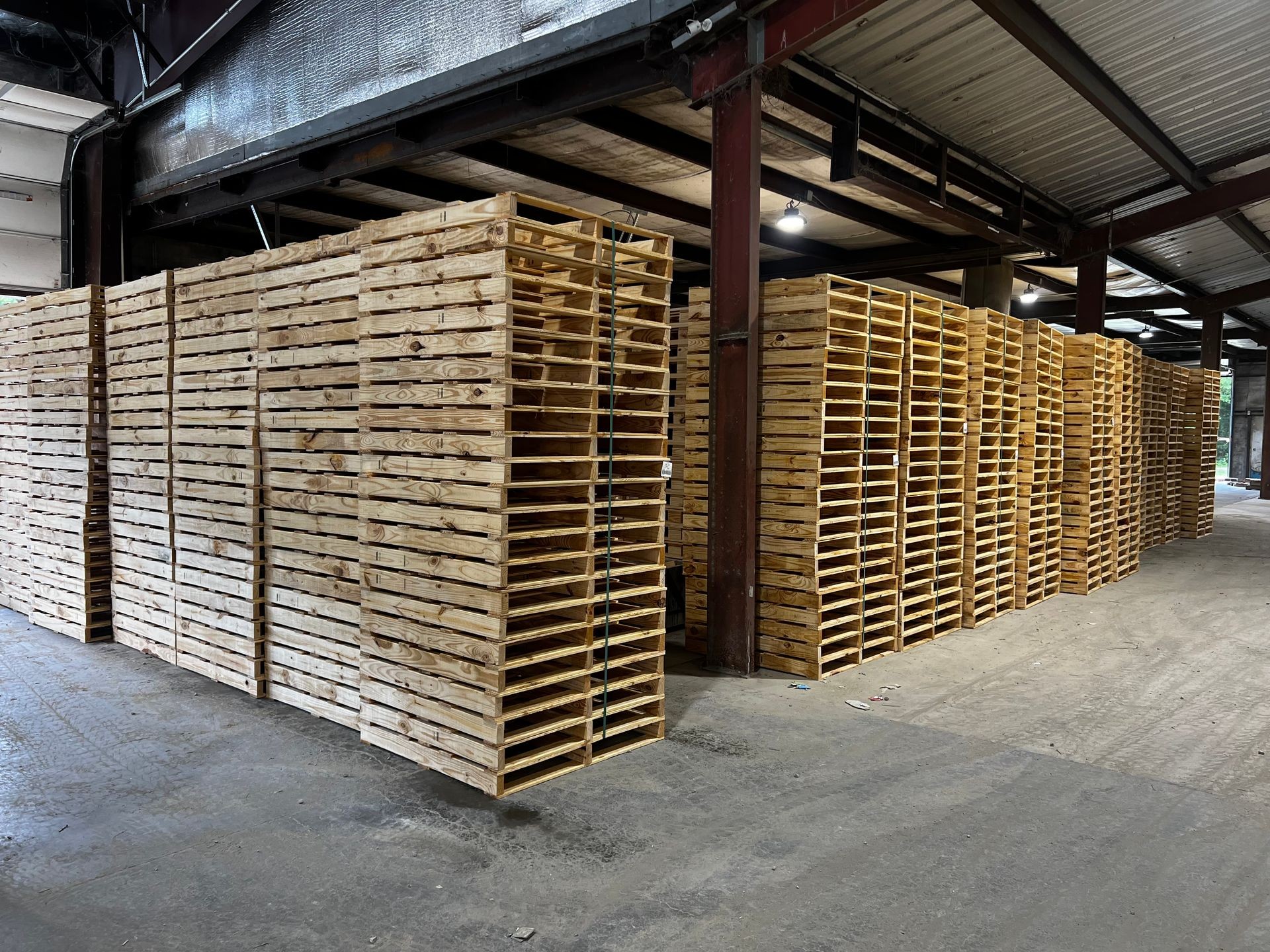 Large stacks of wooden pallets organized in rows inside an industrial warehouse with metal beams and concrete flooring.